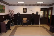 Interior view of a salon area in a senior living facility with two hair dryer chairs, a single chair in the middle, a desk with a computer on the left, a decorative tree, and a wall decal that says 'Love Laugh Live'. The floor is tiled and the walls are painted in two tones with a decorative border near the ceiling.