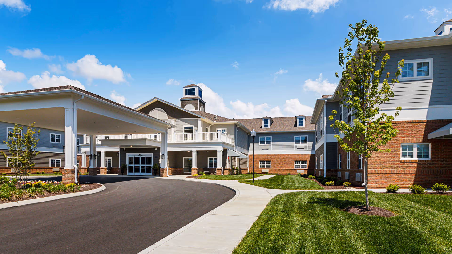 Exterior view of Traditions of Smyrna senior living facility showing a large building with a covered entrance, brick and siding facade, manicured lawn, young trees, and a clear blue sky with some clouds.