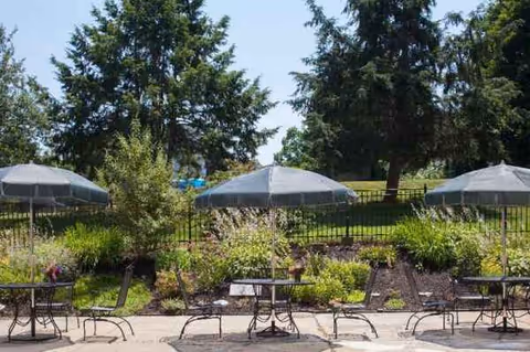 Outdoor patio with metal tables, chairs, and umbrellas amid a landscaped garden.
