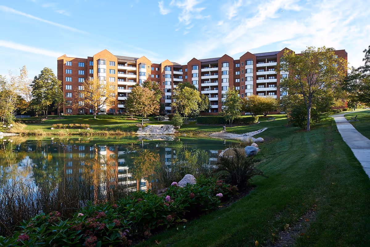 A large multi-story brick building with balconies, surrounded by green trees and landscaping, reflected in a calm pond in the foreground under a blue sky.