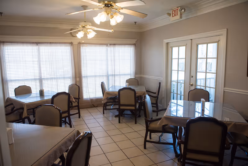 A dining room with multiple tables covered with tablecloths and surrounded by chairs. The room has tiled flooring, large windows with blinds, ceiling fans with lights, and a glass door leading outside.