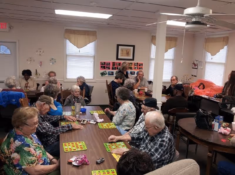 A group of elderly people sitting around tables in a common room, playing bingo and socializing. The room has white walls, windows with beige valances, a ceiling fan, and various decorations on the walls.