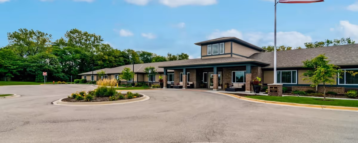 Exterior view of a single-story senior living facility building with a covered entrance, surrounded by a circular driveway and landscaped greenery under a blue sky with some clouds.