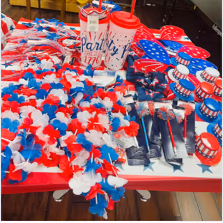 A table covered with a patriotic-themed tablecloth displaying various red, white, and blue party supplies including leis, cups, fans, and small hats, all decorated with stars and stripes.