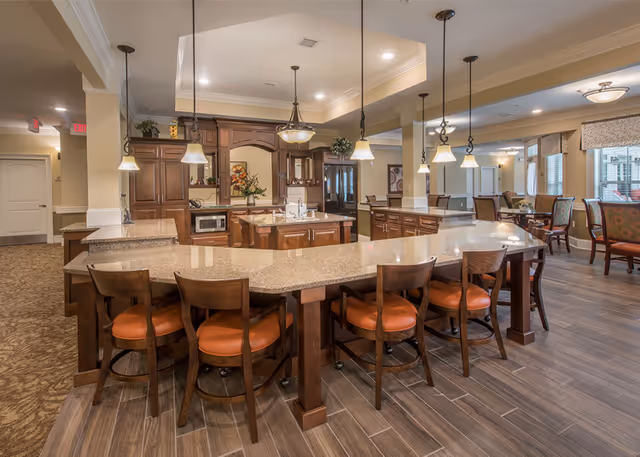 Spacious kitchen and dining area in a senior living facility featuring a large granite countertop island with wooden chairs, pendant lighting, wooden cabinetry, and a seating area with tables and chairs in the background.