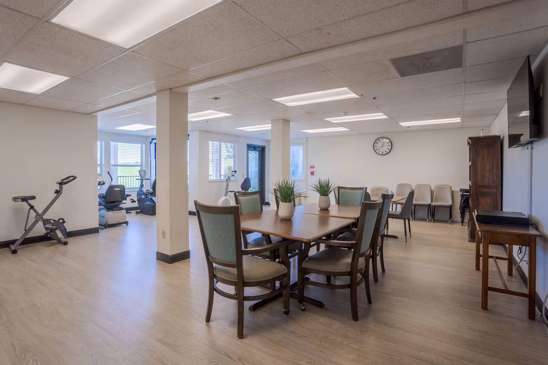 A spacious room with a wooden table surrounded by chairs in the foreground, decorated with small potted plants. In the background, there is exercise equipment near windows letting in natural light. The room has light-colored walls, a clock on the wall, and a television mounted on the right side.
