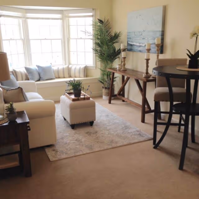A cozy living room with a large window seat adorned with cushions, a white sofa, a small ottoman with a tray and plant on top, a wooden side table with a lamp, a wooden console table with candles and a painting above it, a tall potted plant, and a round dining table with chairs. The room has beige carpet and light-colored walls.