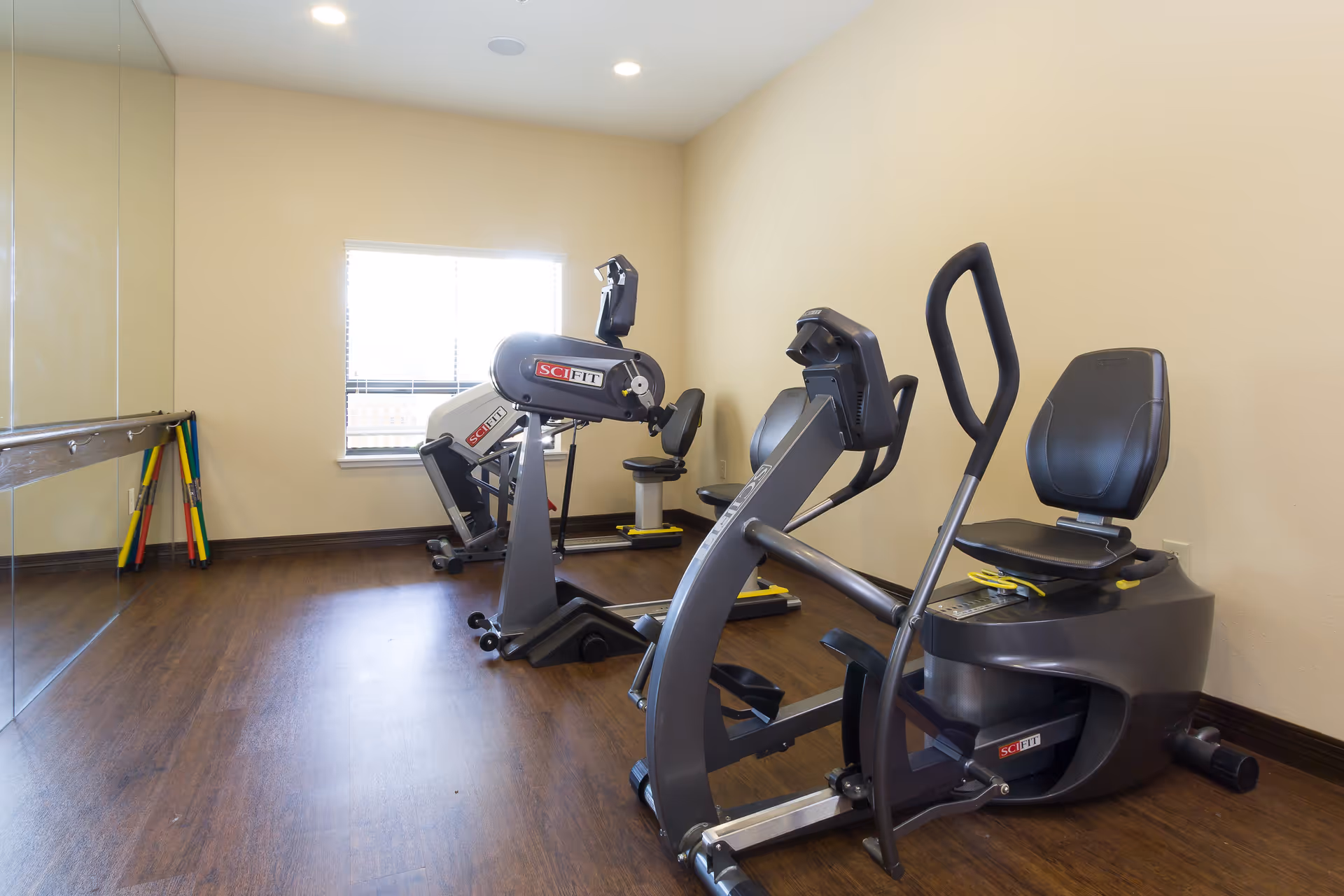 A small exercise room with two SciFit exercise machines positioned on a wooden floor. The room has beige walls, a window with blinds, and a large mirror covering one wall. Some colorful exercise sticks are leaning against the wall near the mirror.