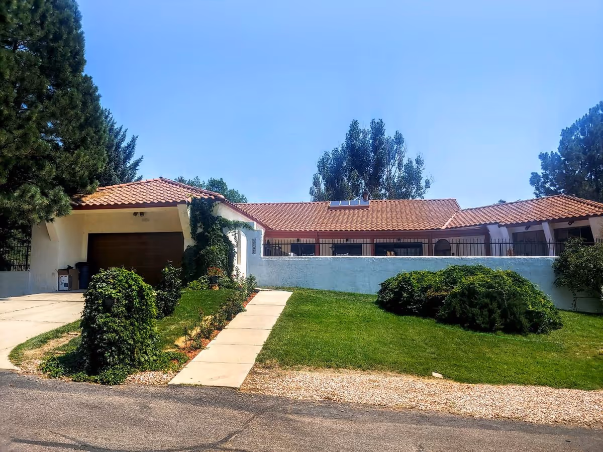 Single-story white stucco building with a red tile roof, garage, front lawn, paved walkway and surrounding trees.