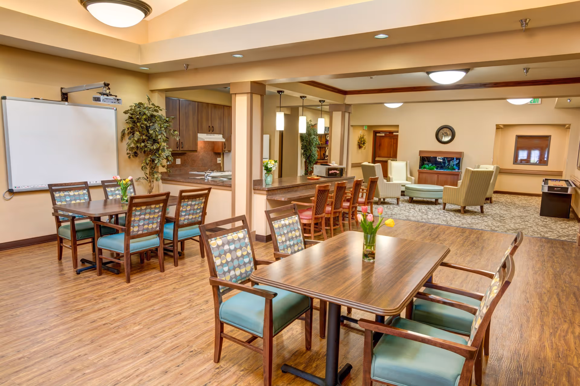 A spacious, well-lit common area in a senior living facility featuring wooden tables with patterned cushioned chairs, a whiteboard on the wall, a kitchen area with bar stools, and a cozy seating area with armchairs and a fish tank in the background.