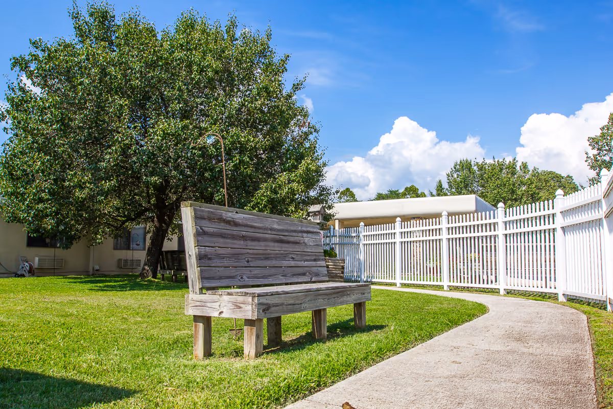 A wooden bench sits on grass beside a curved concrete path and white picket fence with a large tree and blue sky in the background.