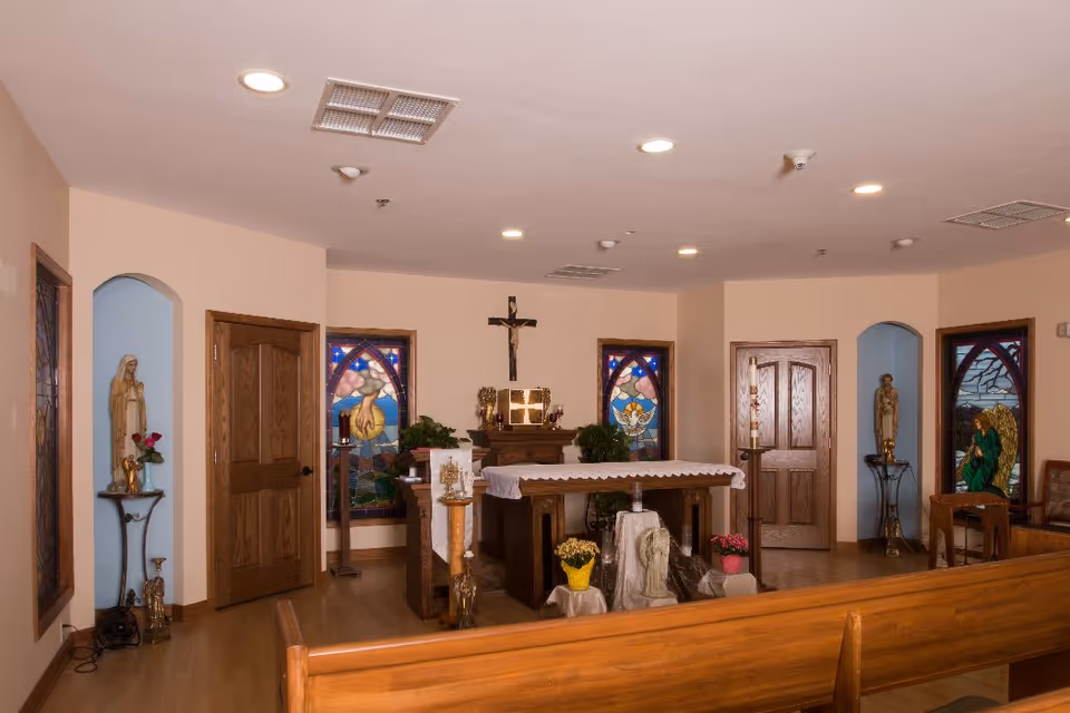 Small chapel interior with wooden pews, an altar topped by a crucifix, stained glass windows, and religious statues.