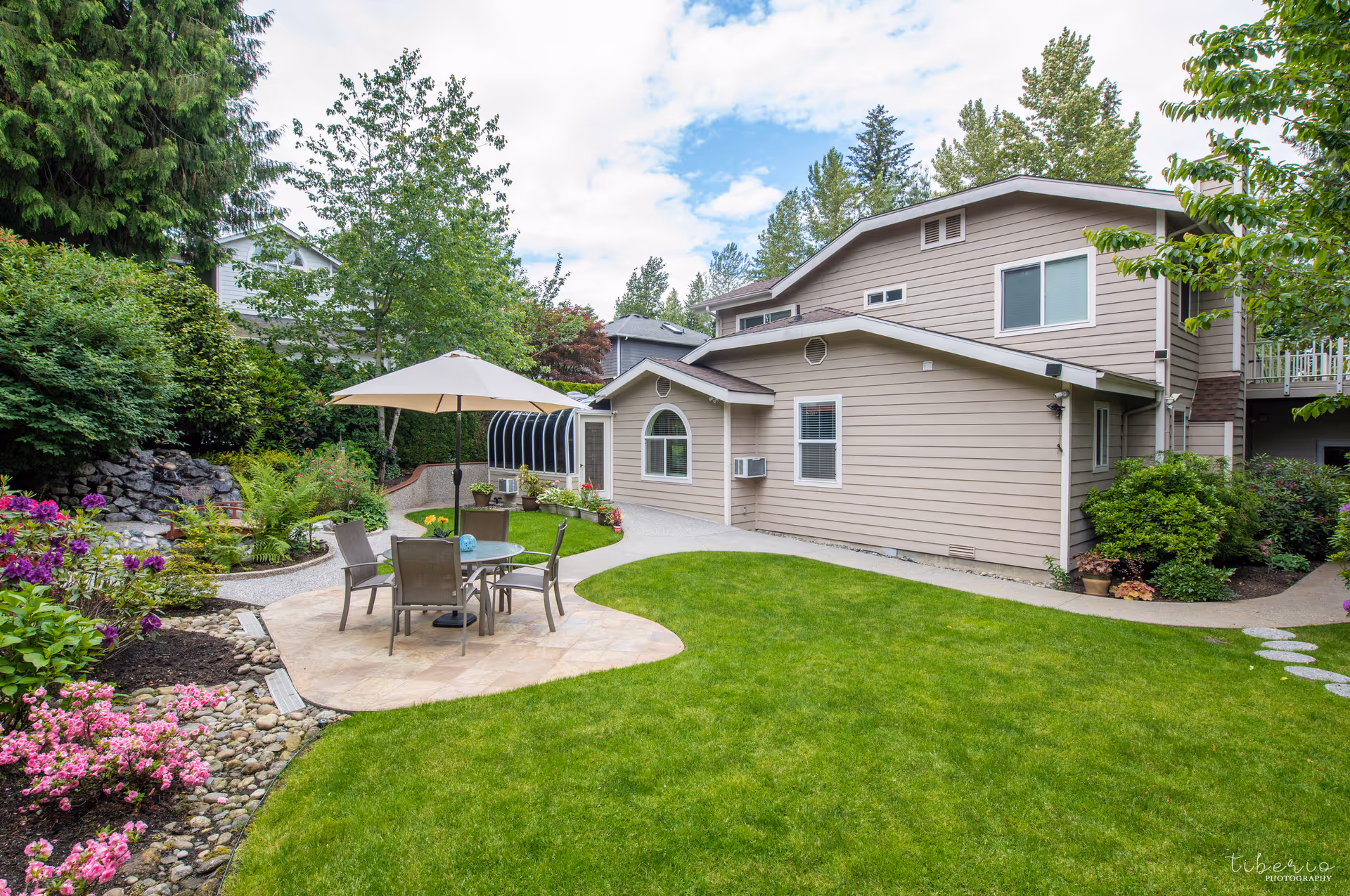 Well-maintained backyard with a patio table and umbrella, green lawn, flower beds, and a beige two-story house exterior.