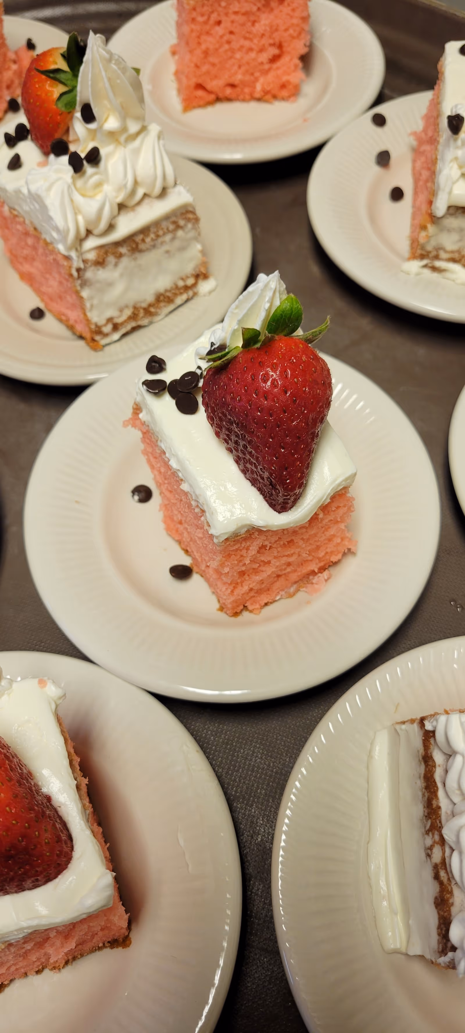 Several small square slices of pink cake with white frosting, topped with a fresh strawberry and chocolate chips, each served on a white plate.