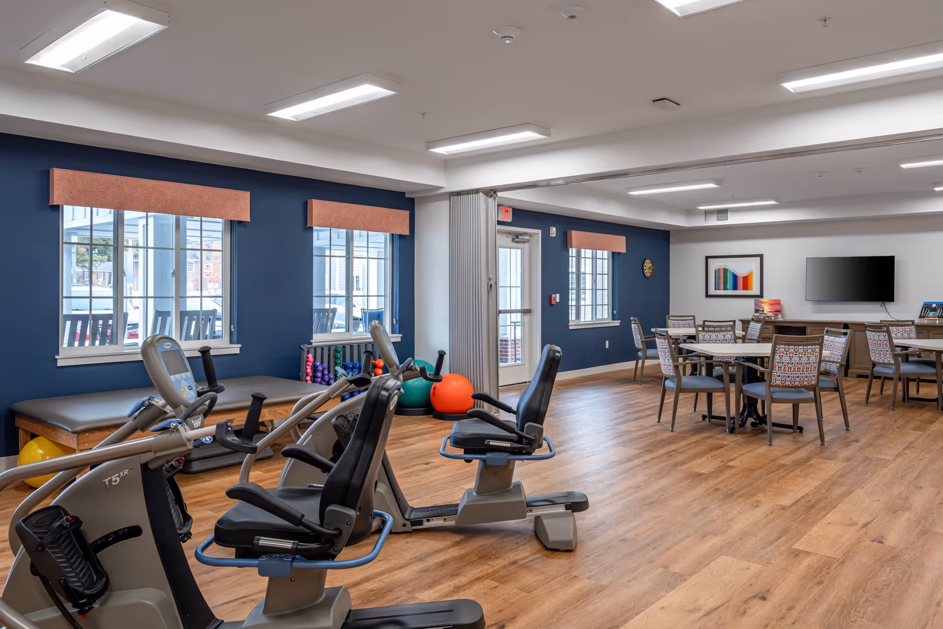 Bright multipurpose senior living activity room with exercise equipment, tables and chairs, and windows along a navy accent wall.