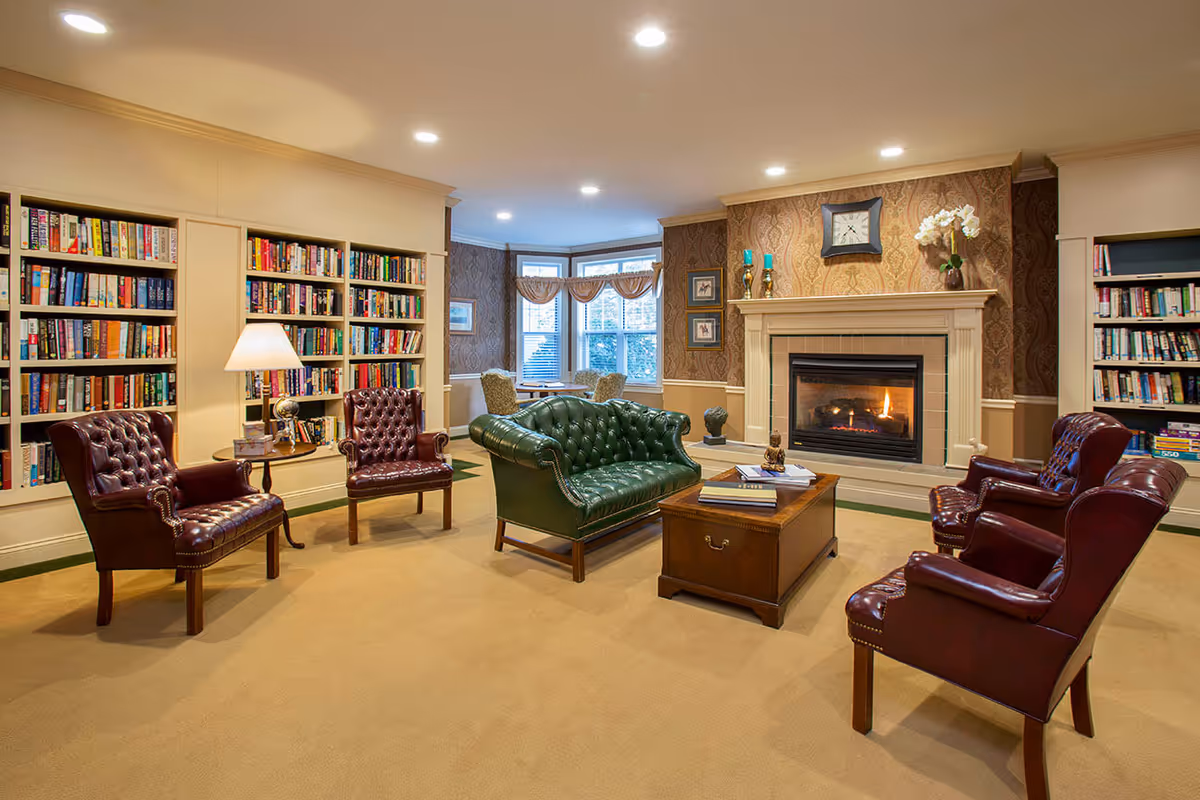A cozy senior living room with a green leather sofa and four burgundy leather armchairs arranged around a wooden coffee table. The room features built-in bookshelves filled with books, a lit fireplace with decorative items on the mantel, and a bay window with two chairs and a small table. The walls have patterned wallpaper and the floor is carpeted in beige.