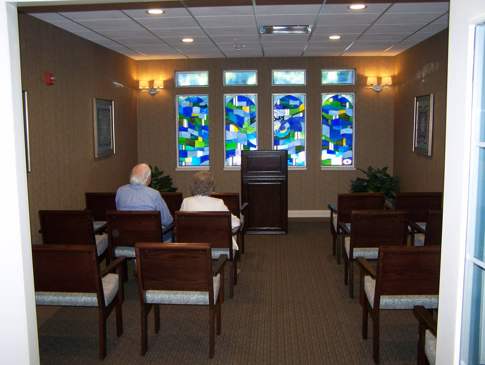 A small room with rows of wooden chairs with cushioned seats facing a wooden podium. Two elderly people are seated in the front row. The back wall features four stained glass windows with abstract blue, green, and yellow designs. The room has beige walls, carpeted floor, and wall-mounted light fixtures.
