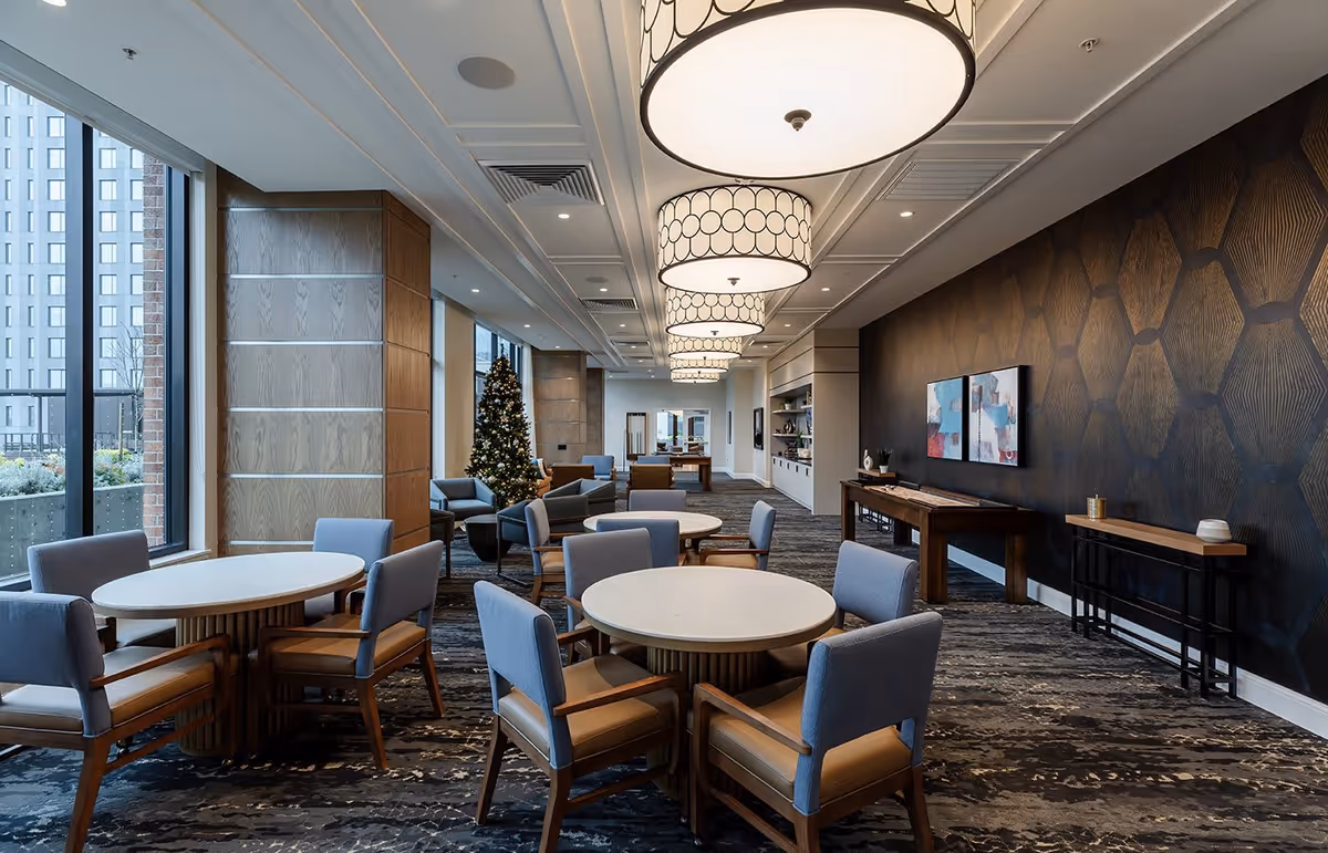 A spacious, modern common area with round tables and chairs arranged for seating. Large windows on the left side let in natural light and show an outdoor view of buildings. The room features a patterned dark accent wall with artwork, multiple large circular ceiling lights, and a decorated Christmas tree near the windows. There are additional seating areas and tables further back in the room.