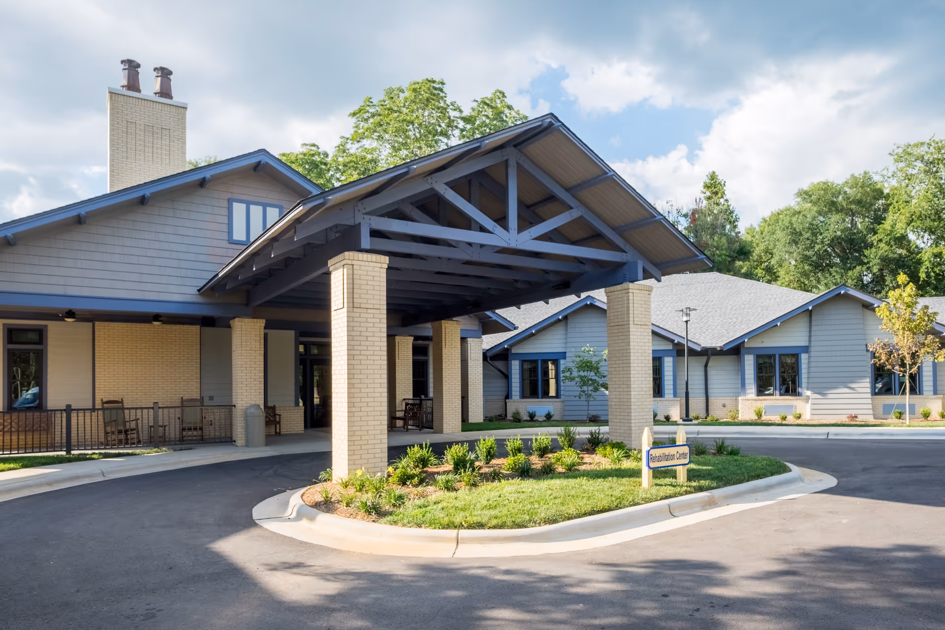 Exterior view of White Oak Manor Shelby showing a covered entrance supported by brick pillars, a circular driveway, and a sign indicating the Rehabilitation Center. The building has light-colored brick and siding with blue trim, surrounded by greenery and trees under a partly cloudy sky.