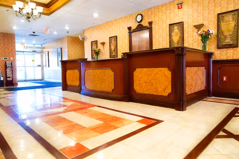 A spacious reception area with a large wooden front desk featuring ornate paneling. The floor has a decorative tile pattern in shades of cream and orange. The walls are covered with patterned wallpaper and decorated with framed artwork and flower arrangements. A chandelier hangs from the ceiling, and there is a clock mounted on the wall behind the desk. The entrance doors with glass panels are visible in the background.