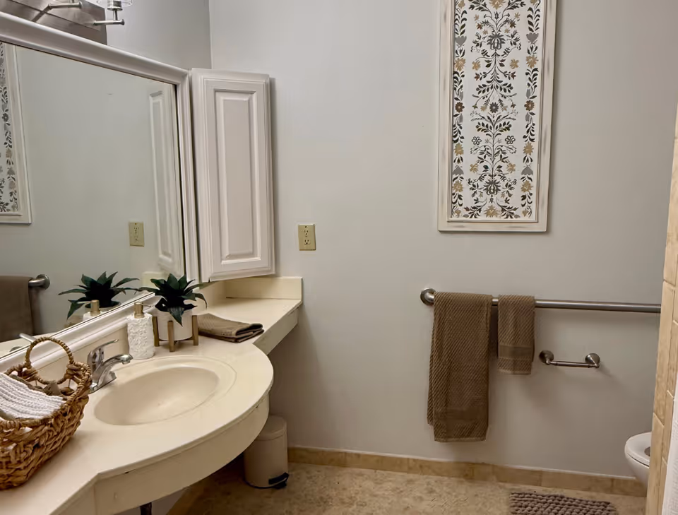 A clean, accessible bathroom with a curved sink countertop, mirror, towel bars and framed wall art.