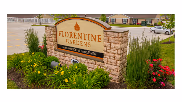 Stone sign for Florentine Gardens Transitional Care & Rehabilitation surrounded by green plants and flowers with a parking lot and residential buildings in the background.