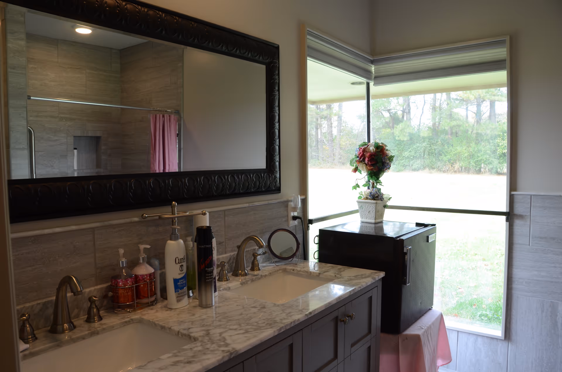 Bathroom interior with a double sink vanity featuring marble countertops, two faucets, and various toiletries including soap dispensers and lotion. A large decorative mirror hangs above the sinks. In the background, a shower with a pink curtain is visible. To the right, there is a window with a view of greenery outside, and a small black refrigerator topped with a flower arrangement in a white pot.
