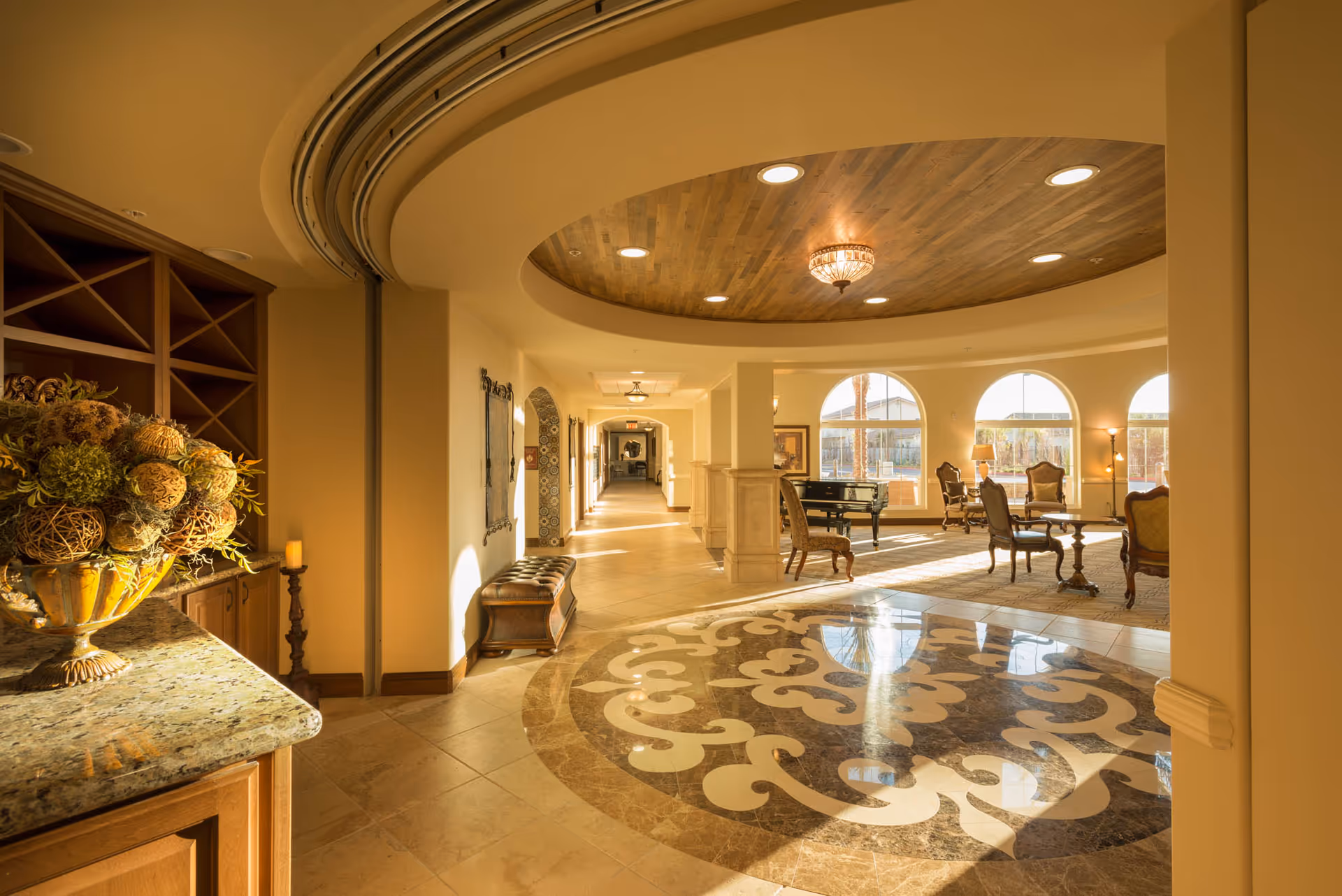 A spacious and elegant senior living facility interior with a decorative tiled floor, arched windows letting in natural light, classic wooden furniture including chairs and tables, a piano, and a ceiling with recessed lighting and a chandelier.