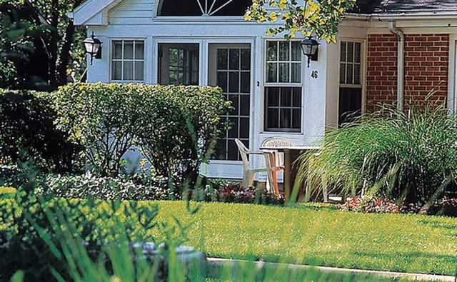 A well-maintained outdoor garden area in front of a building entrance with a white door and windows. There are two outdoor chairs and a small table on a porch surrounded by green bushes, grass, and flowering plants.