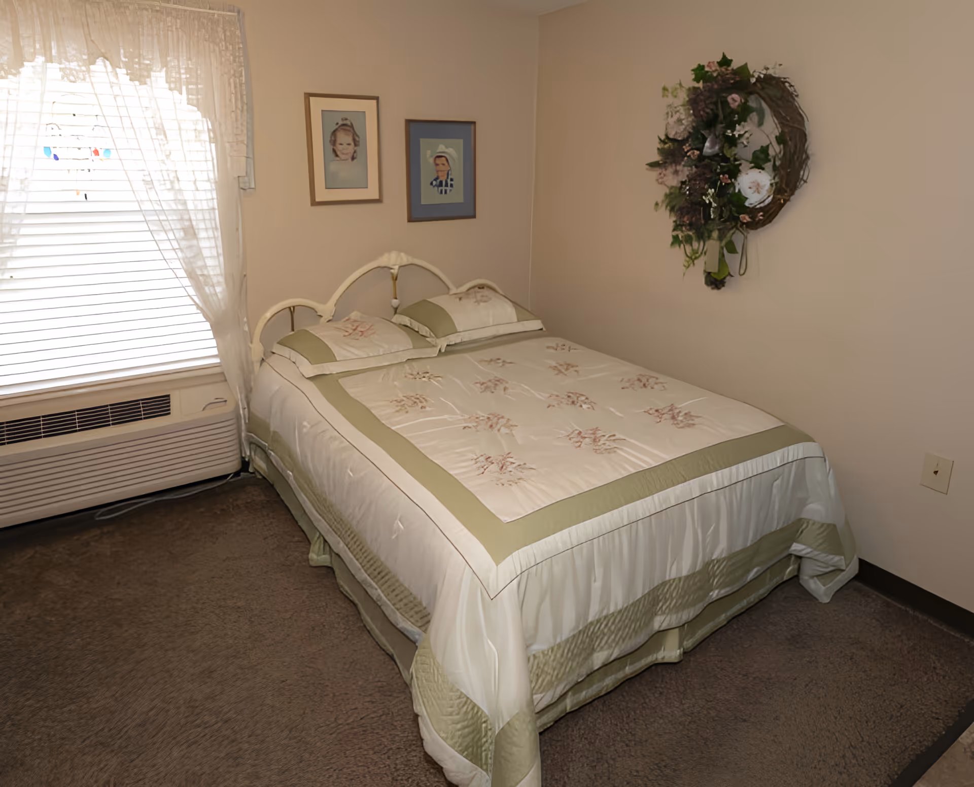 Small bedroom with a double bed dressed in green-and-white floral bedding, a window with lace curtains, framed portraits on the wall, and a decorative wreath.