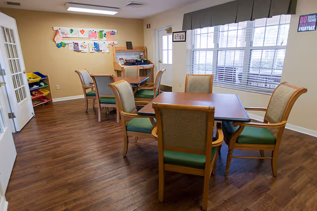 A well-lit room with wooden flooring featuring a square table surrounded by six cushioned chairs with wooden frames. The room has large windows with blinds on one side, a bulletin board with colorful artwork on the wall, and a small desk with a chair in the corner. There is also a shelving unit with colorful trays near the door.