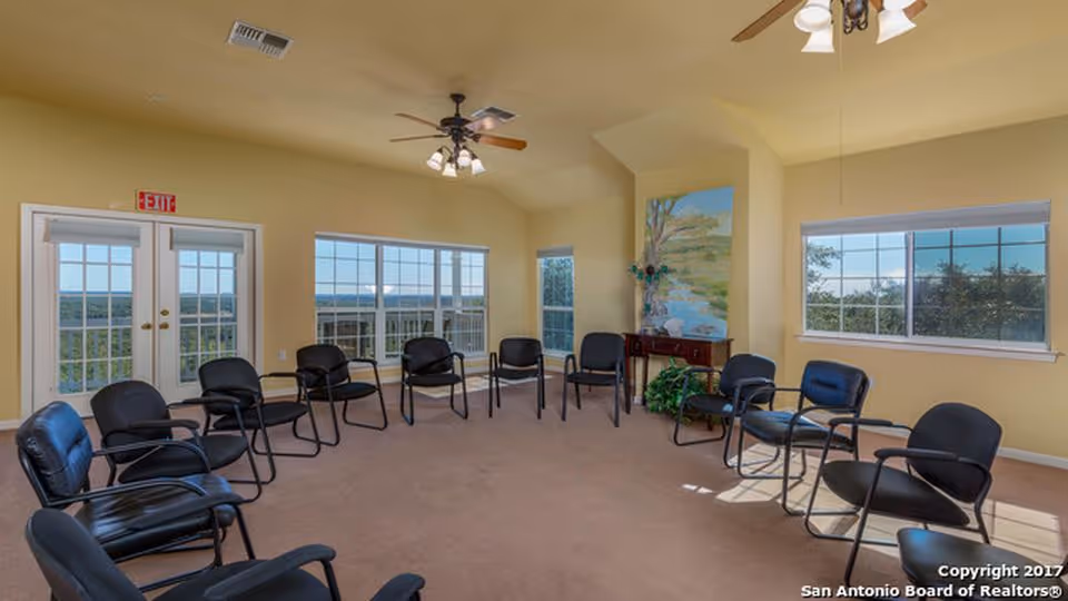 A spacious room with beige walls and carpeted floor, arranged with black chairs in a large circle. The room has multiple windows letting in natural light, a ceiling fan with lights, and a small table with a plant and a painting on the wall. Double doors with glass panes lead outside.