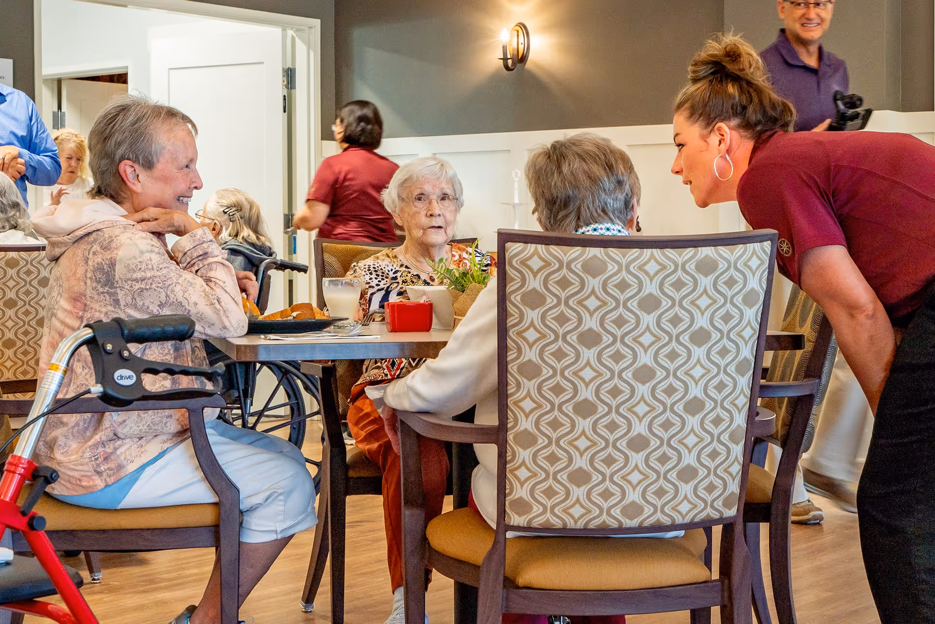 A group of elderly women sitting around a dining table in a senior living facility, engaging in conversation. A staff member is leaning in to talk with one of the women. The room has wooden flooring, patterned chairs, and a warm, inviting atmosphere.