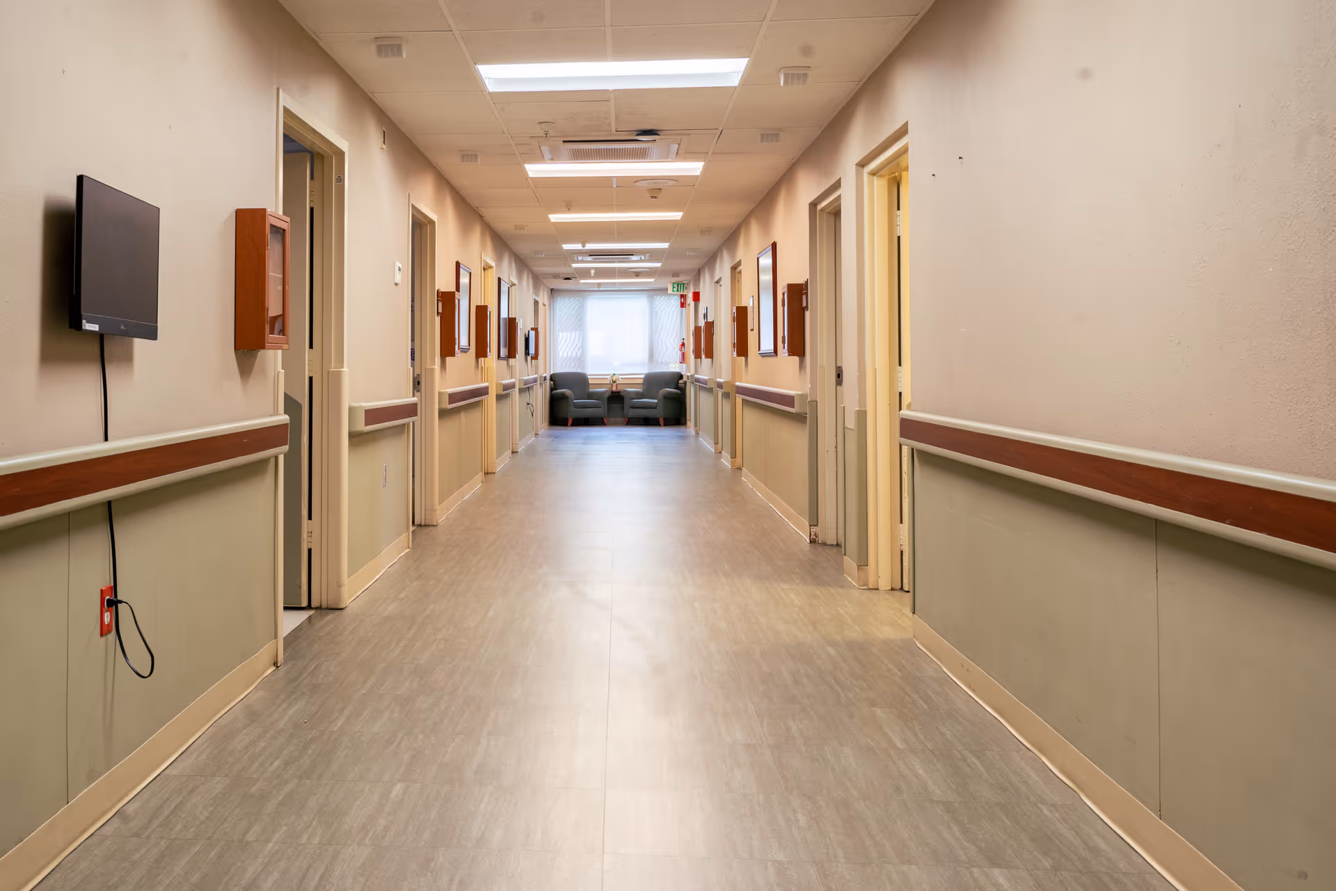 A long, empty hallway in a healthcare facility with beige and light green walls, several open doorways on both sides, handrails along the walls, a wall-mounted TV on the left, and a seating area with two armchairs and a small table at the far end near a window.