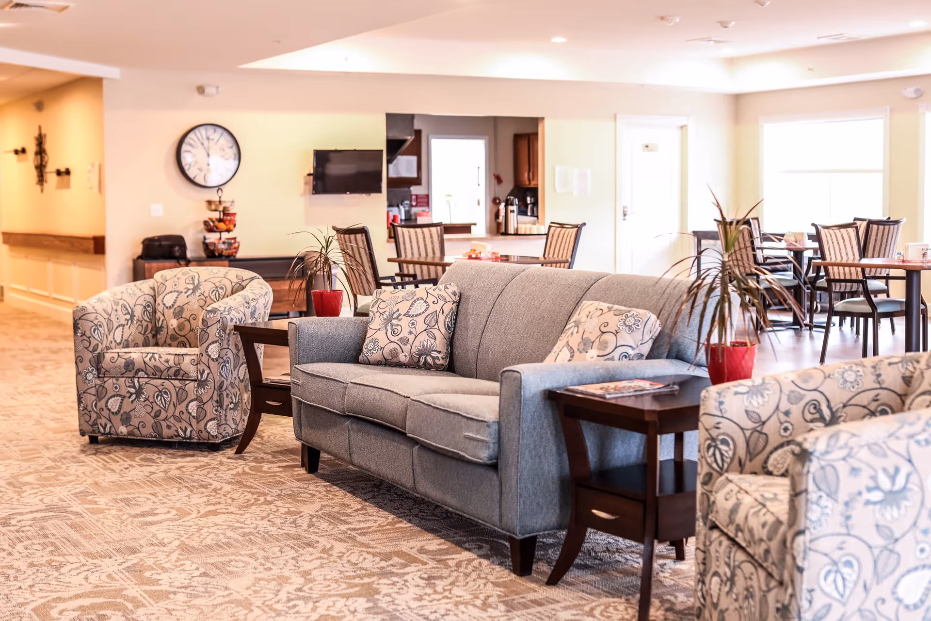 A bright and spacious common area in an assisted living facility featuring a gray sofa with patterned cushions, two matching armchairs with floral designs, wooden side tables with red potted plants, and a dining area with tables and chairs in the background. A wall clock and a mounted TV are visible on the far wall.
