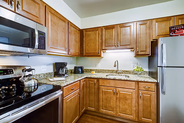 A modern kitchen with wooden cabinets, granite countertops, a stainless steel microwave, stove, and refrigerator. The countertop holds a coffee maker, toaster, a glass container with green apples, and a small dish with a yellow item. The kitchen has under-cabinet lighting and a stainless steel sink with a faucet.