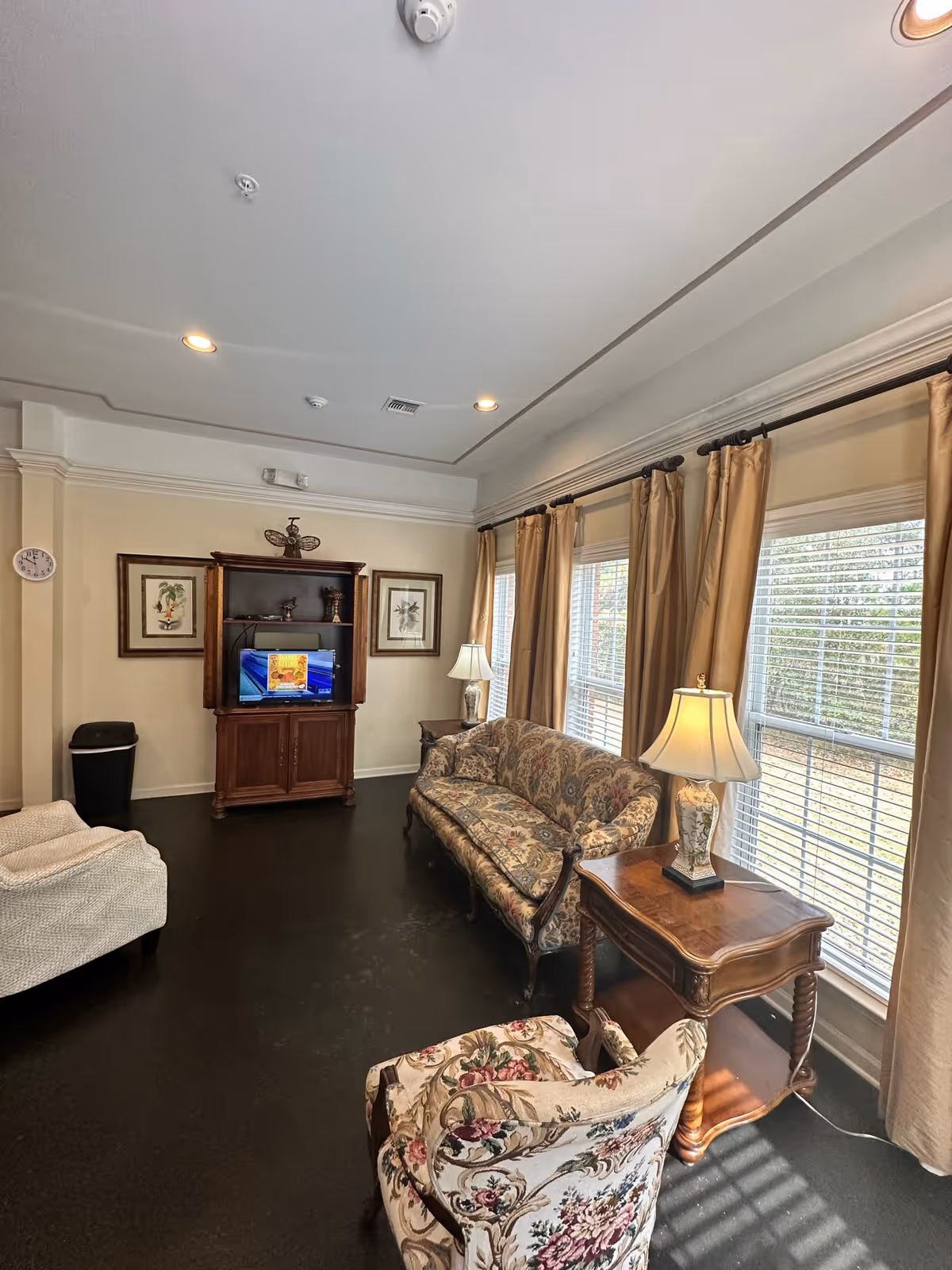 A furnished living room with patterned sofa and chairs, a wooden TV cabinet, side tables with lamps, and large curtained windows letting in sunlight.