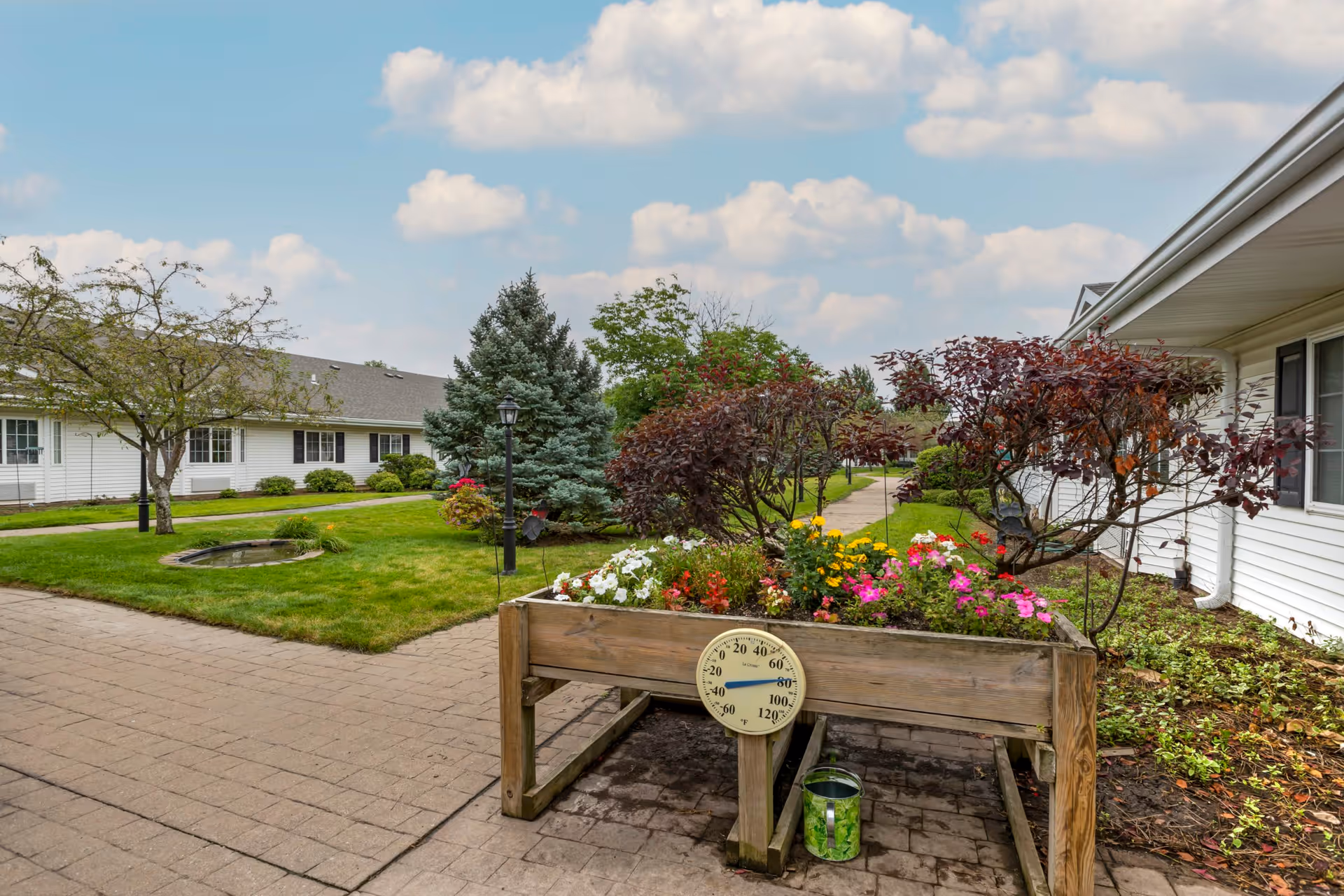 Courtyard of a senior living facility with a raised wooden flower planter and thermometer, paved walkways, lawn, trees, and white single-story buildings.