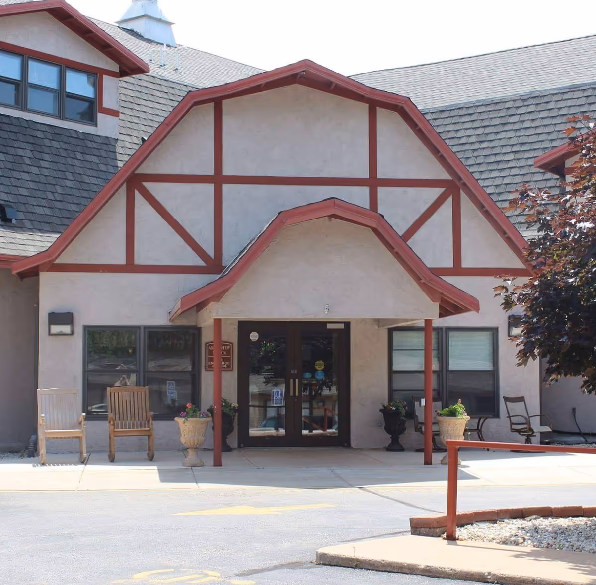 Front entrance of Arborview Manor, a building with beige walls and red trim. The entrance has double glass doors under a peaked roof supported by red posts. There are wooden chairs and potted plants on either side of the entrance, with a tree visible on the right side.