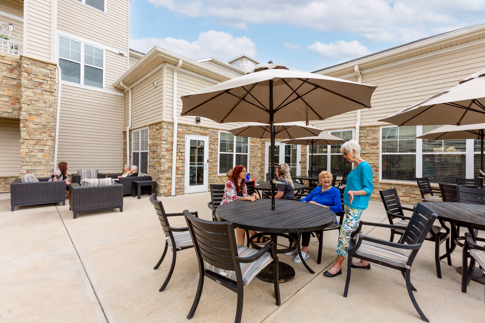 Outdoor patio area at StoryPoint Troy with several round tables and chairs under large umbrellas. Elderly women are sitting and standing, engaging in conversation. The building exterior features beige siding and stone accents with large windows and doors.