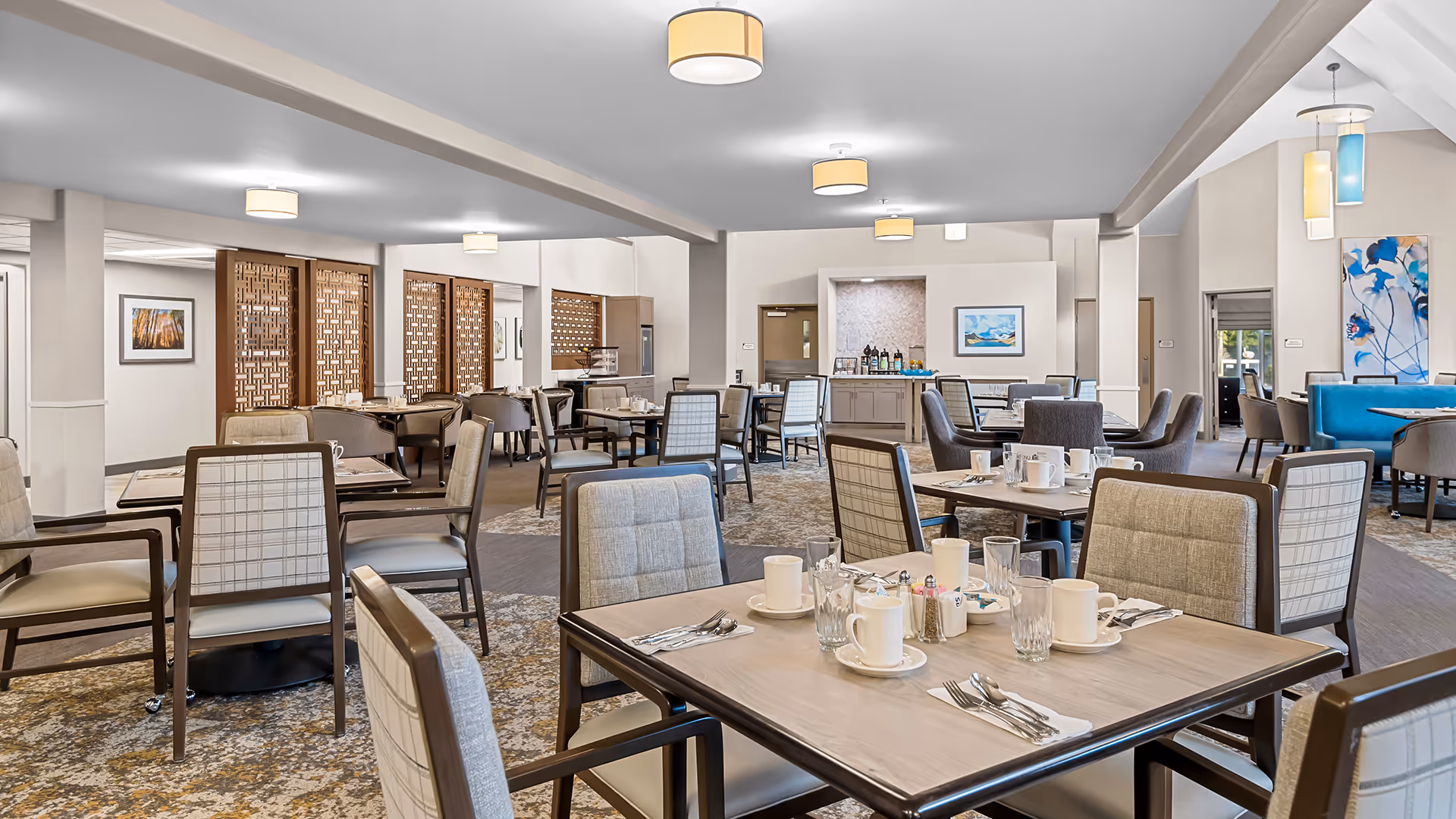 A spacious, well-lit dining room with multiple tables and chairs arranged neatly. Each table is set with white cups, glasses, utensils, and napkins. The room features modern light fixtures on the ceiling, decorative wooden partitions, framed artwork on the walls, and a carpeted floor with a patterned design. There is a counter area in the back with beverages and snacks.