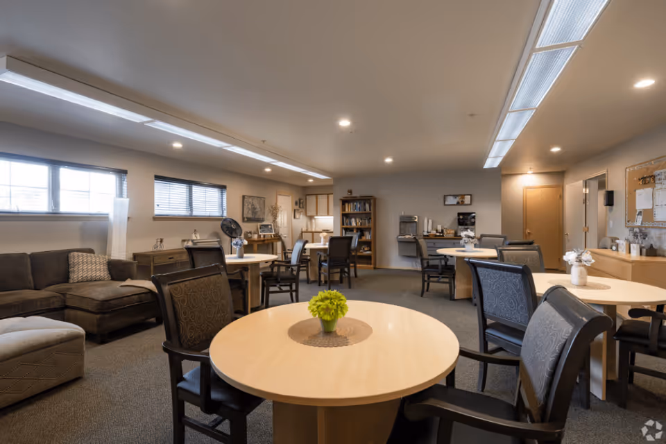 A spacious common area in a senior living facility with round tables and chairs arranged for socializing or activities. There is a brown sectional sofa and ottoman on the left side, a bookshelf and a water dispenser against the far wall, and soft overhead lighting illuminating the room.