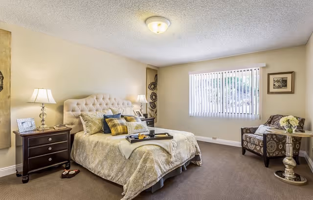 A well-lit bedroom with a large bed featuring a tufted headboard, multiple pillows, and a tray on top. There are two bedside tables with lamps and picture frames. A comfortable armchair and a small round table with a flower arrangement are placed near a window with vertical blinds. The room has beige walls and carpeted flooring.
