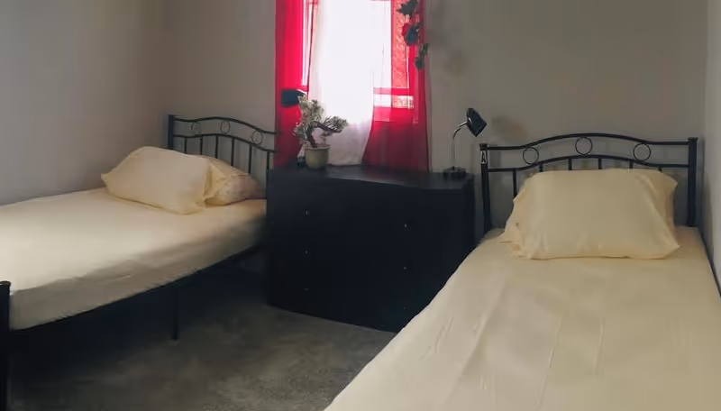 Small bedroom with two twin beds separated by a dark dresser beneath a window with red curtains.