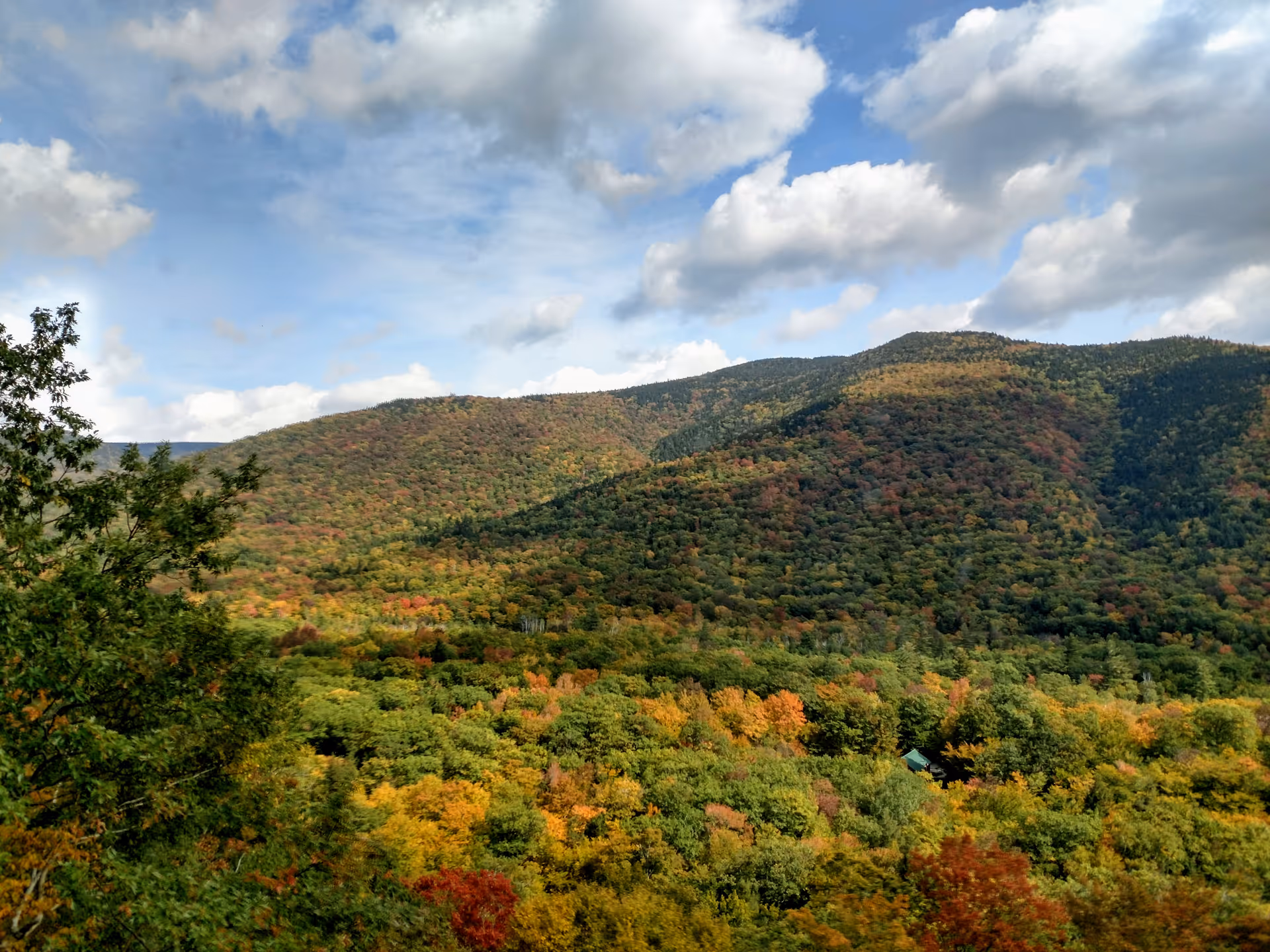 View of forested mountains with autumn foliage under a partly cloudy sky.