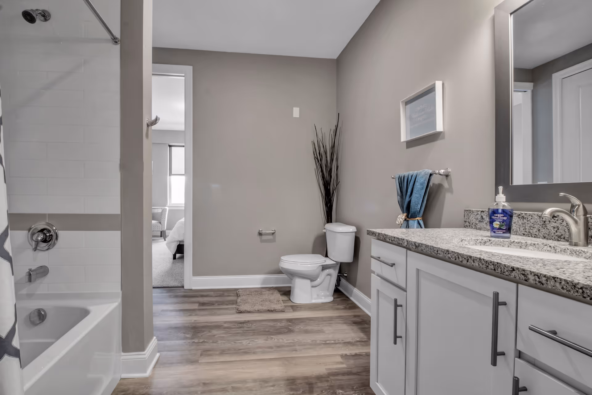 A modern bathroom with a white bathtub and shower on the left, a toilet in the center against a beige wall, and a white vanity with a granite countertop and a large mirror on the right. A blue towel hangs on a rack above the toilet, and a bottle of liquid hand soap is on the countertop. The bathroom has wood-style flooring and a doorway leading to a bedroom with a window and chair visible.