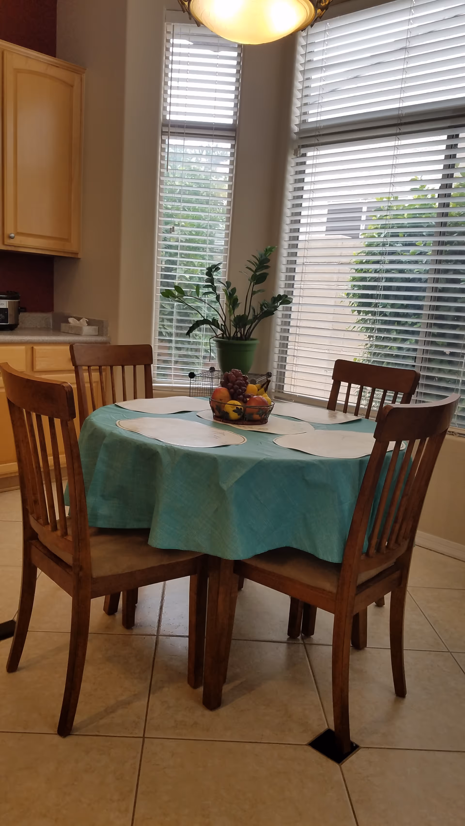 A dining area with a round wooden table covered with a turquoise tablecloth and four wooden chairs. The table has placemats and a fruit basket centerpiece. Behind the table are large windows with white blinds and a potted plant on the windowsill. Part of a kitchen counter and cabinets are visible to the left.