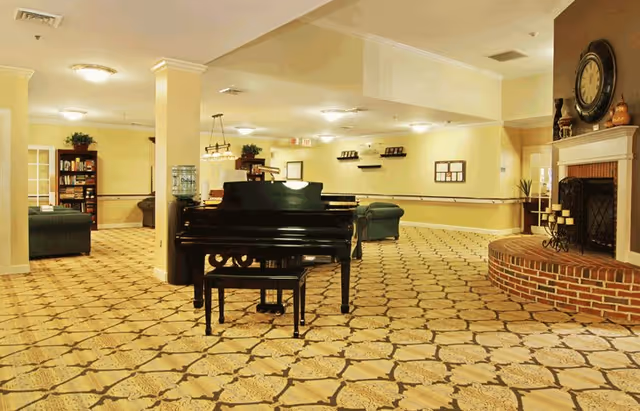 A spacious senior living common area featuring a black grand piano with a matching bench in the center, patterned carpet flooring, a brick fireplace with candles and a large clock above it, and comfortable seating areas with green sofas. The room is softly lit with ceiling lights and decorated with plants and framed pictures on the walls.