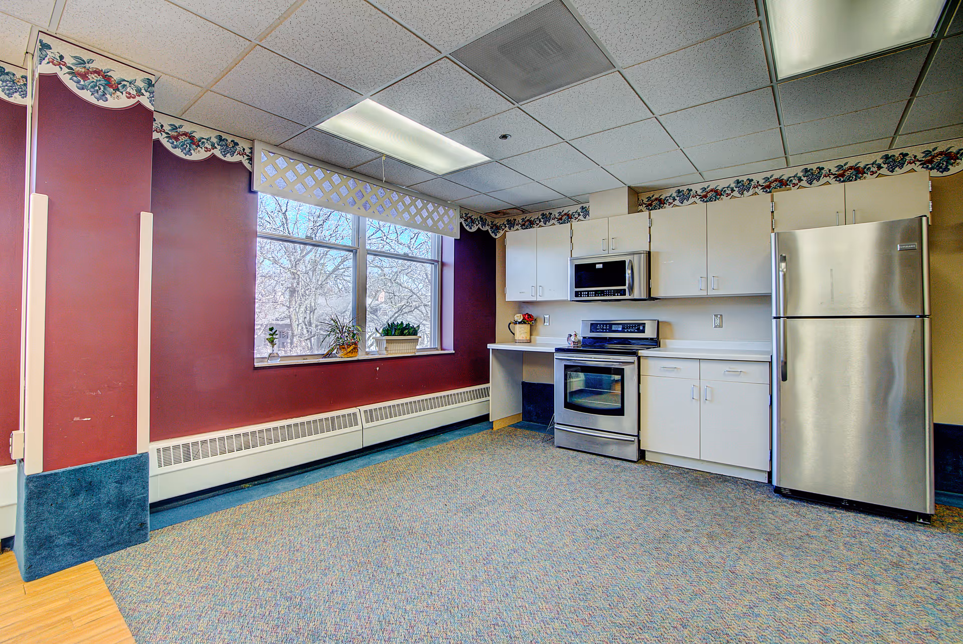 A bright communal kitchenette with stainless steel appliances, white cabinets, and a large window overlooking trees.