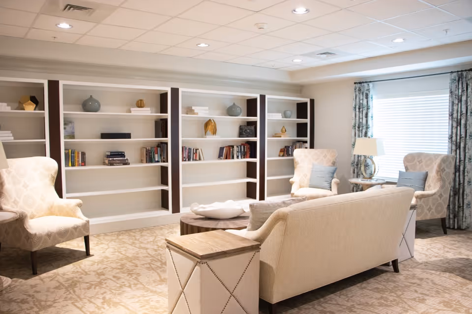 A bright and cozy living room with beige upholstered chairs and a sofa arranged around a wooden coffee table. Behind the seating area is a large built-in bookshelf with decorative items and books. A window with patterned curtains allows natural light into the room.